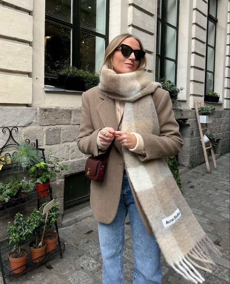 Woman in stylish winter outfit with scarf, sunglasses, and coat standing by a rustic urban backdrop.
