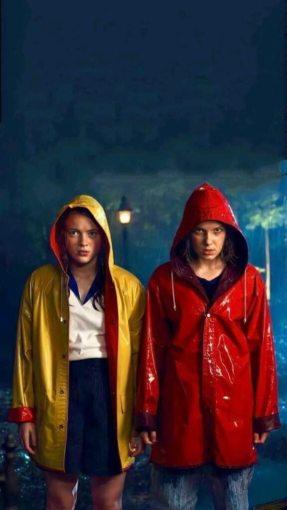 Two people in yellow and red raincoats standing in the rain at night with a street lamp in the background.