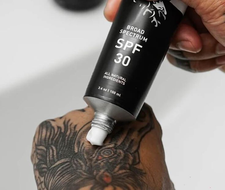 Tattooed hand applying SPF 30 sunscreen from a black tube with natural ingredients.