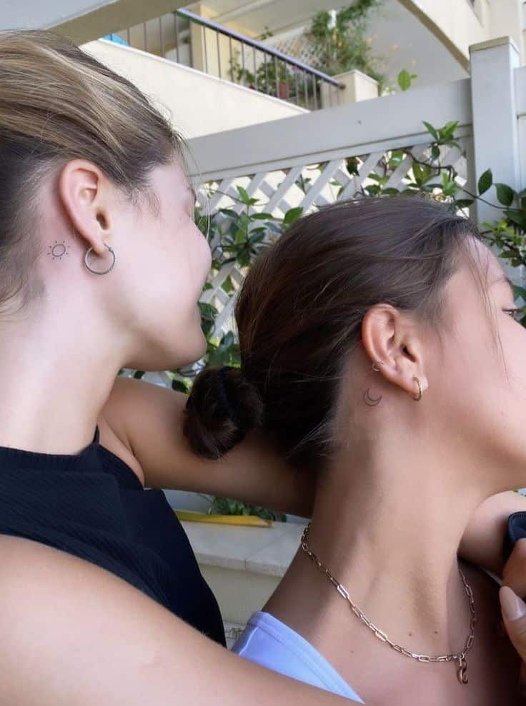 Two women with matching sun and moon tattoos behind their ears, smiling outdoors with greenery in the background.