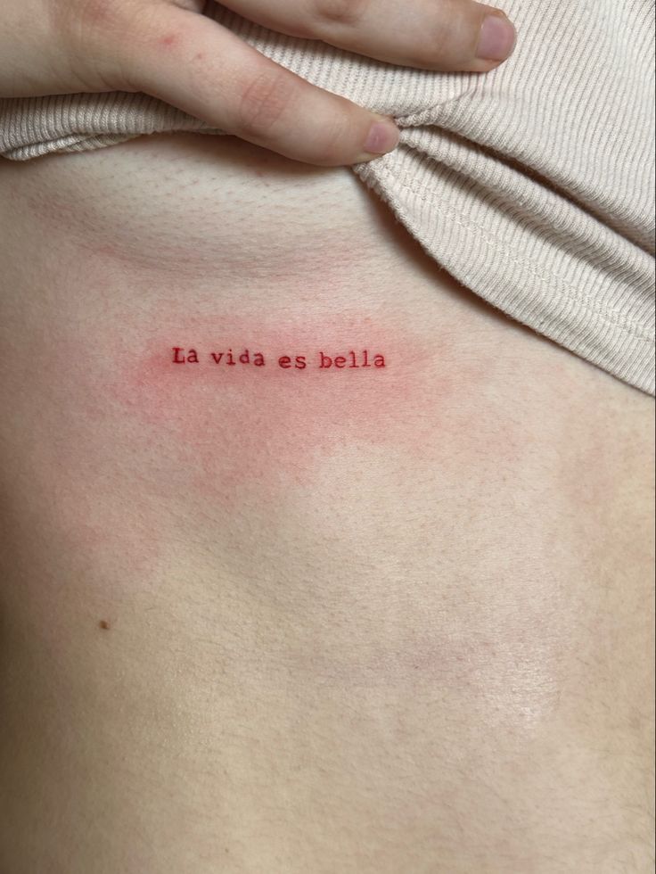 Minimalist red text tattoo 'La vida es bella' on skin, emphasized by lifted beige shirt.