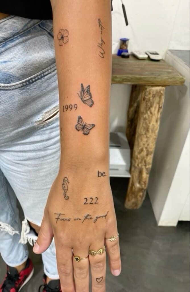 Arm with various minimalist tattoos including butterflies, text, and symbols. Casual jeans with rings on fingers.