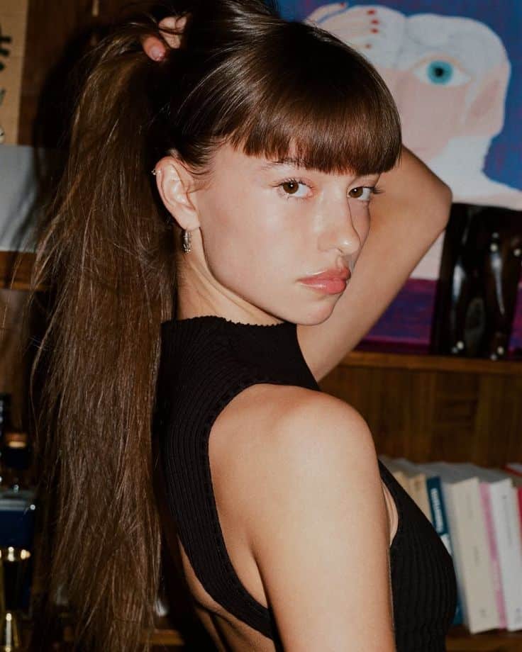 Young woman with long hair and bangs in a black top looks over shoulder, posing indoors with books on a shelf behind her.