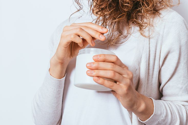 Person applying moisturizer from a jar, wearing a white sweater, promoting skincare and wellness.