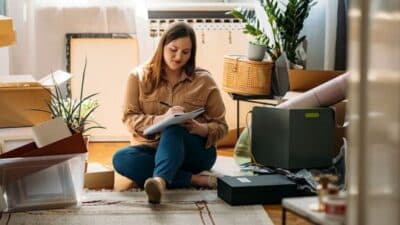 Woman seated among boxes, writing in a notebook. Moving or organizing in a cozy, plant-filled room.