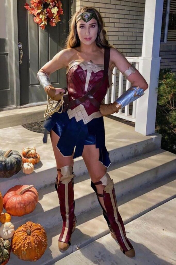 Woman in Wonder Woman costume on porch with pumpkins, Halloween decor in background.