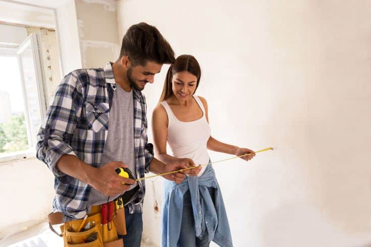 Couple measuring a wall for home renovation, smiling and using a tape measure in a bright, unfinished room.