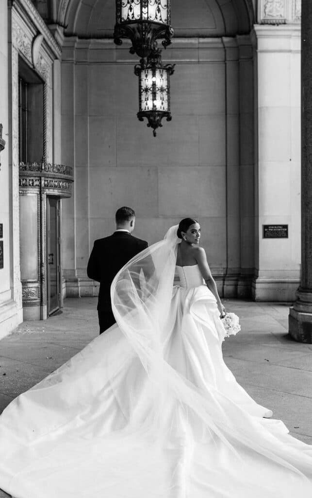 Elegant bride in flowing gown with groom, back view, under ornate chandelier in grand venue. Black and white portrait.