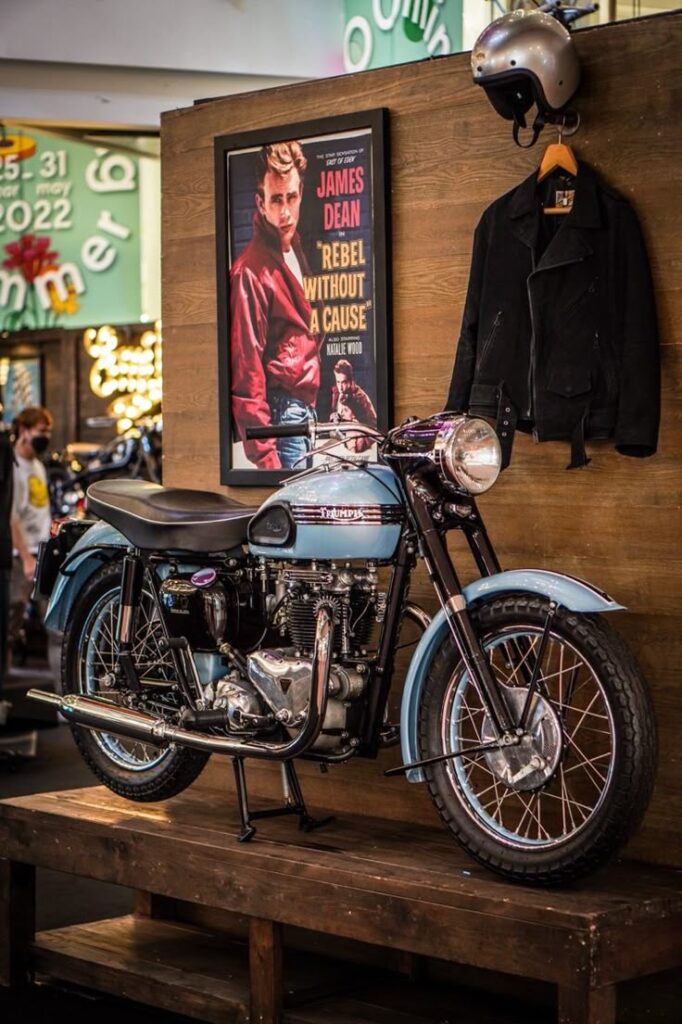 Vintage Triumph motorcycle with leather jacket and helmet, James Dean poster in background. Classic 1950s style display.
