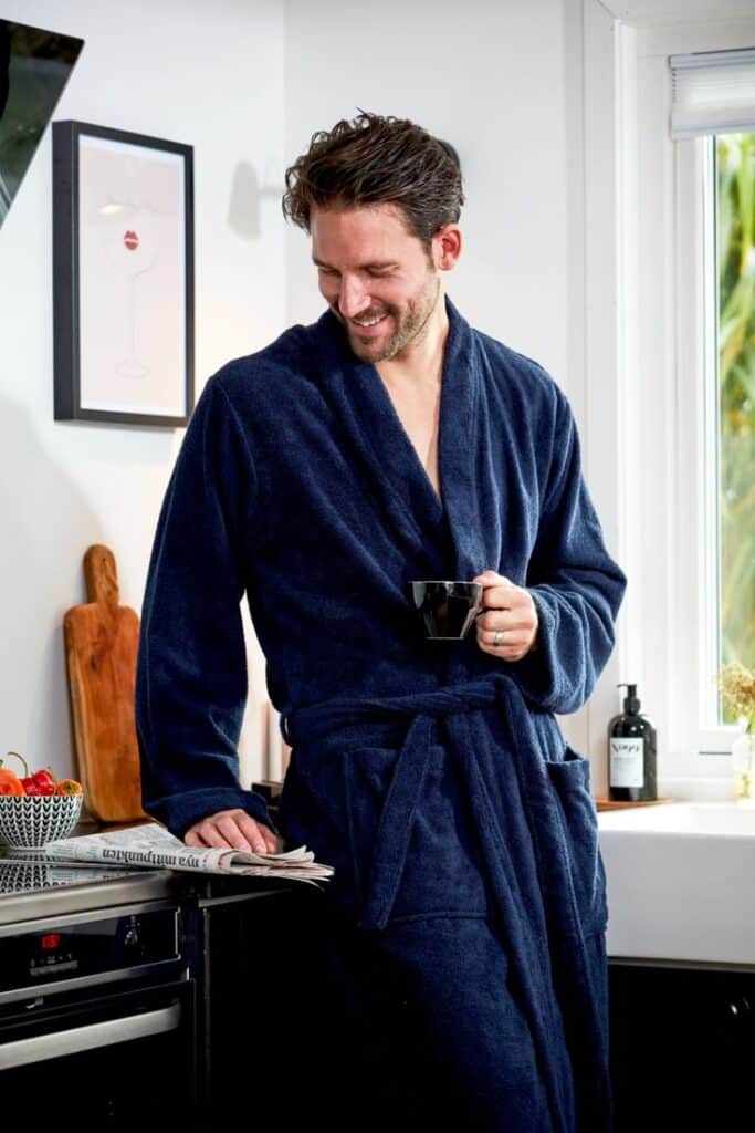 Man in a navy robe enjoys coffee in a cozy kitchen setting, smiling while holding a newspaper.