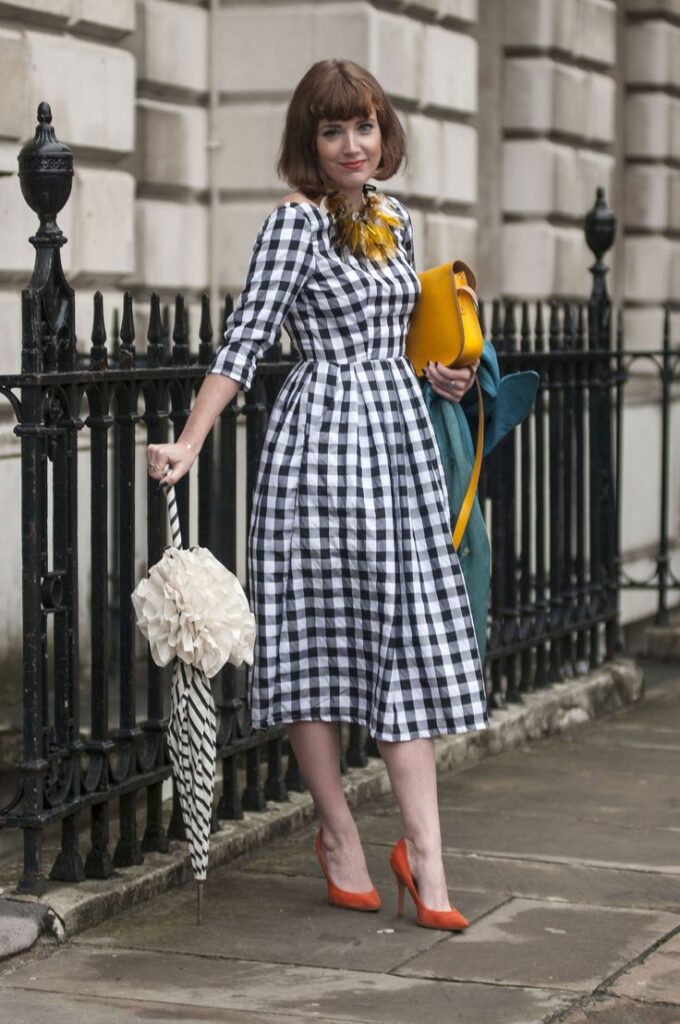 Stylish woman in a gingham dress holding a yellow clutch and a decorative umbrella, standing by a wrought iron fence.