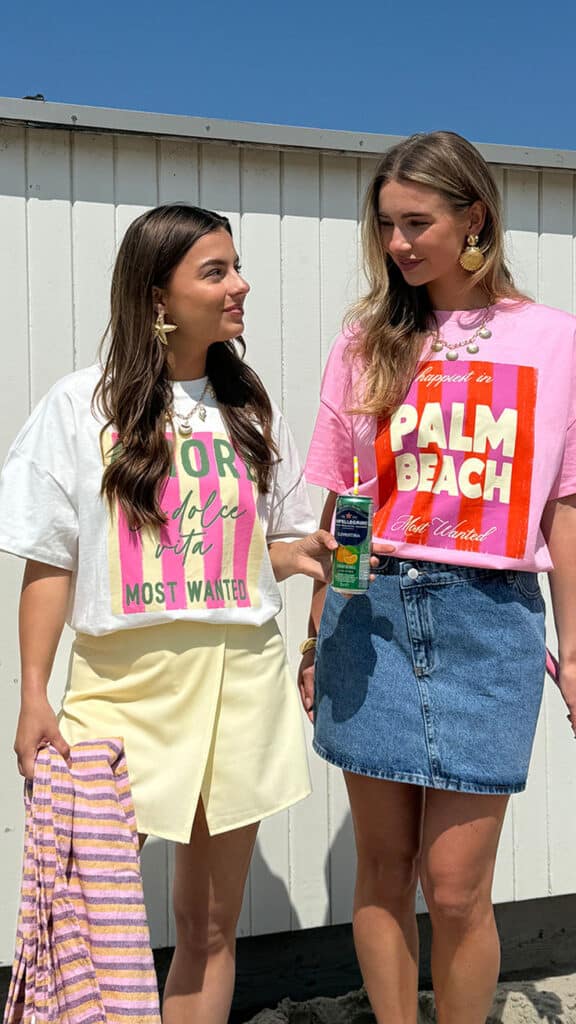 Two women enjoying summer beach style in graphic tees, denim skirt, and holding a drink, under blue skies.