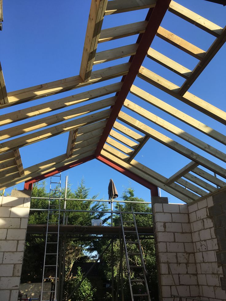 Wooden roof frame construction with clear blue sky background, featuring scaffolding and ladders.