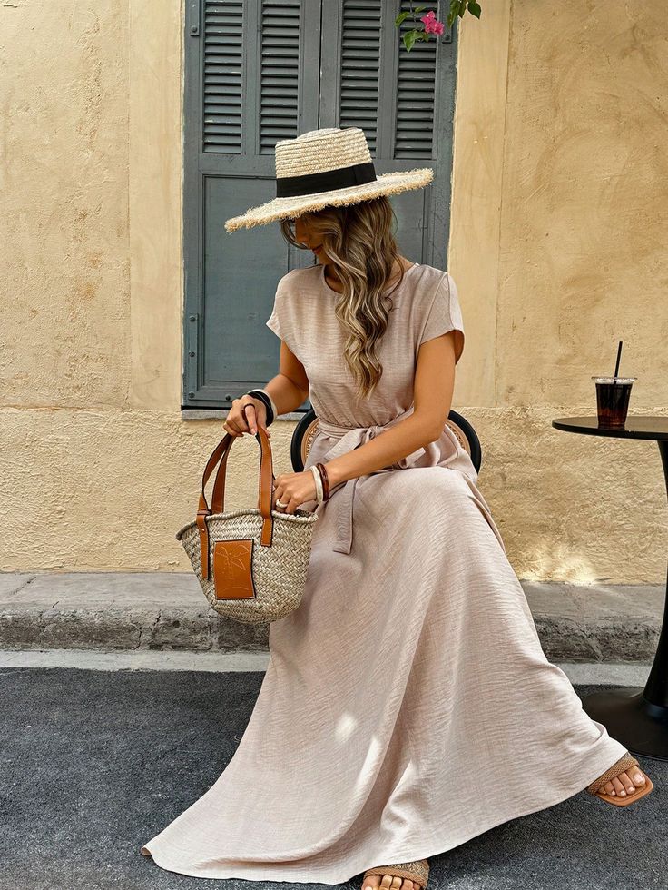 Woman in straw hat and beige dress sitting outside, holding a wicker bag, with a drink on a small table.
