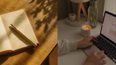 Open notebook with a pen on a sunlit desk and hands typing on a laptop near a lit candle on a cozy workspace.