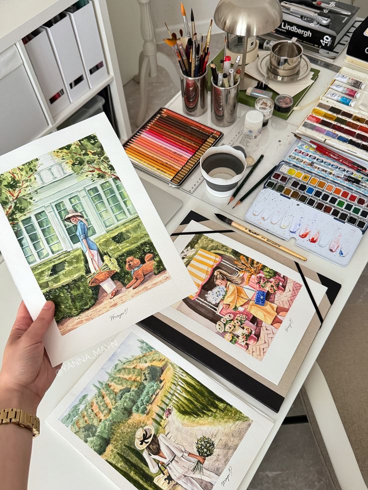 Art studio desk with colorful paints, brushes, and three bright watercolor paintings of scenic views.