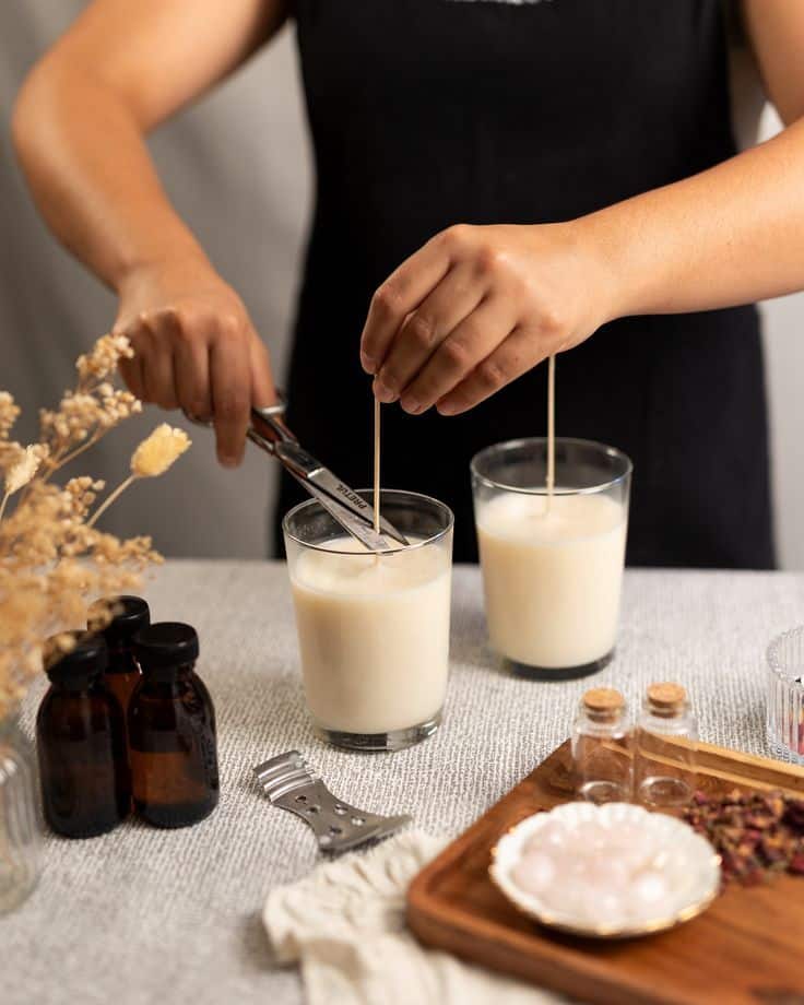 Person crafting homemade candles with wicks in glass jars, surrounded by essential oils and natural ingredients.