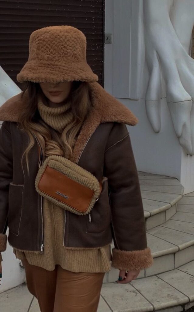 Stylish woman in cozy brown faux fur coat and hat with leather bag, standing on stairs.