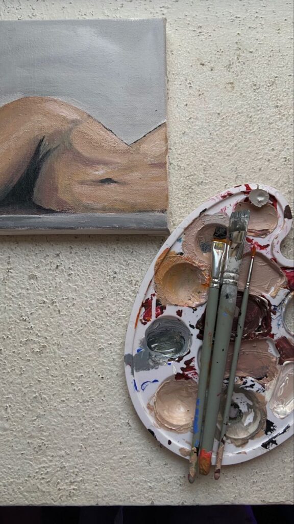 Painting palette with brushes next to a nude art canvas on a textured surface.