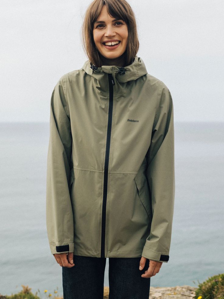 Smiling person in green hooded jacket outdoors near ocean.