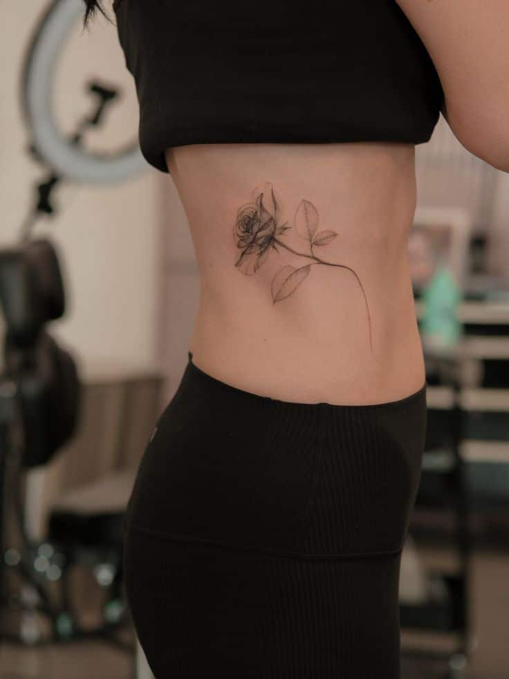 Woman displaying a detailed rose tattoo on side torso, wearing black clothing in a studio environment.
