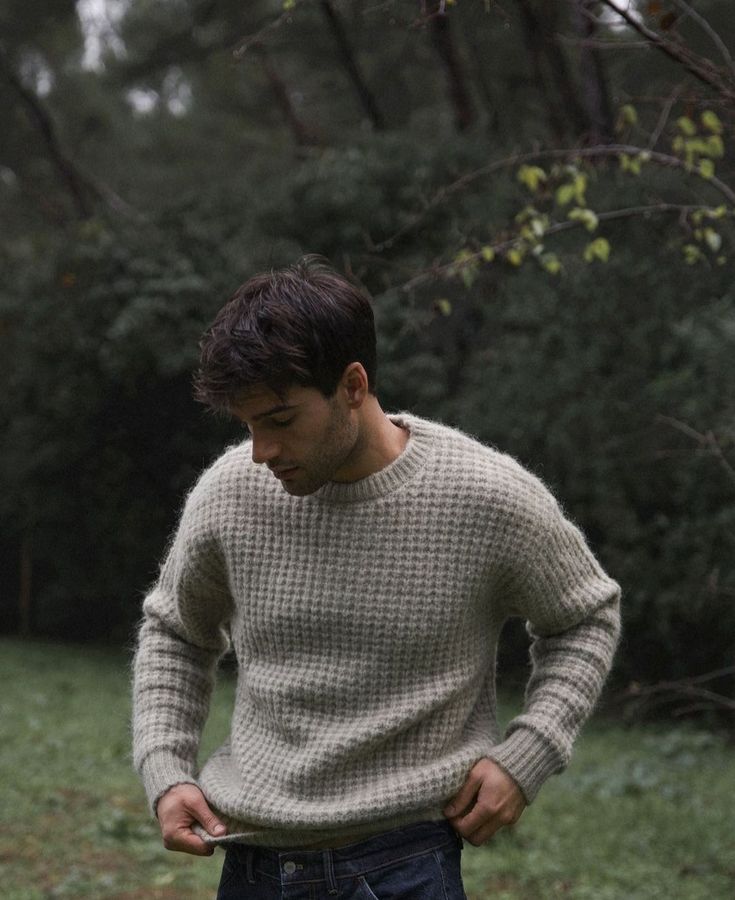 Man in a cozy sweater standing in a forest setting, looking down thoughtfully.