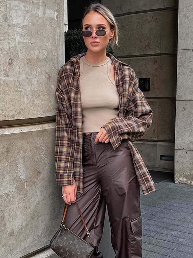 Fashionable woman in sunglasses wearing a plaid jacket and cargo pants, holding a designer bag outdoors.