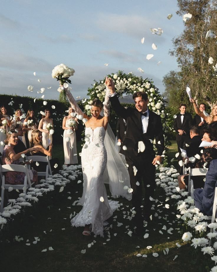 Bride and groom celebrate under floral arch with guests cheering and tossing petals at outdoor wedding.