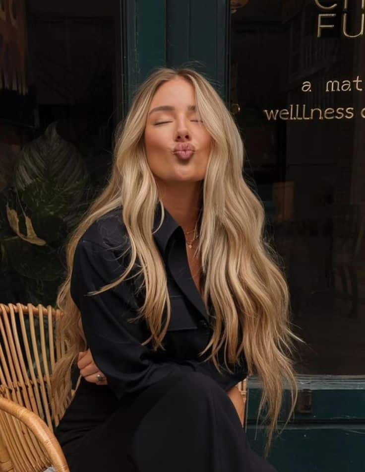 Woman with long blonde hair blows a kiss, wearing a black outfit. Cozy cafe background with greenery.