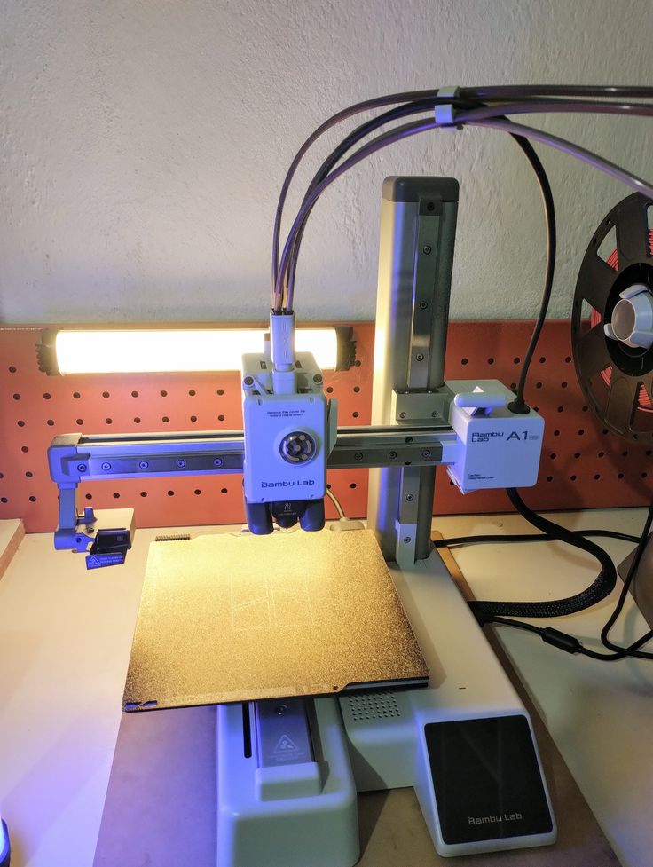 Desktop 3D printer in a workshop with a lit work bed and pegboard background.
