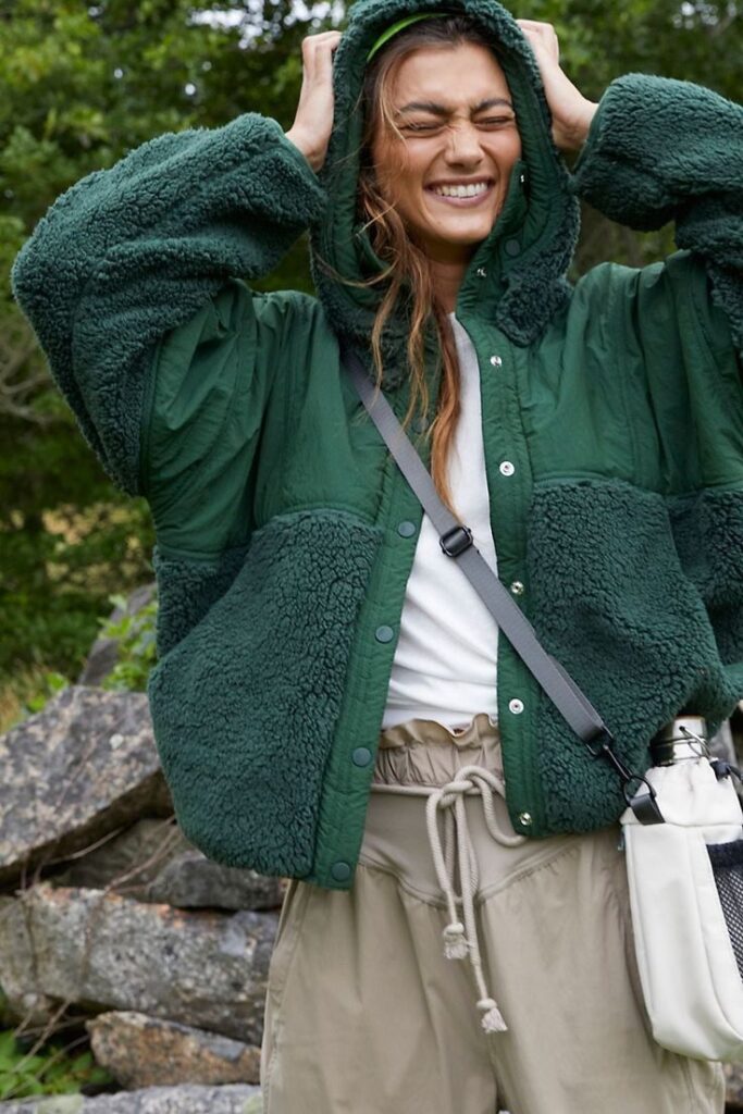 Woman in green fleece jacket smiles outdoors, standing amidst rocky landscape and trees.