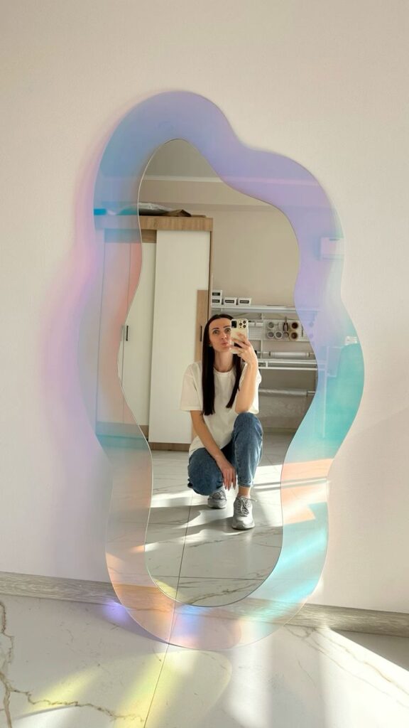 Woman taking a selfie in front of a colorful, wavy mirror, wearing a casual outfit and smiling.