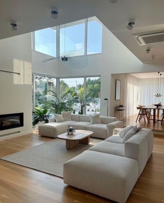 Modern living room with large windows, white sofas, wooden floor, and dining area, featuring natural light and greenery.