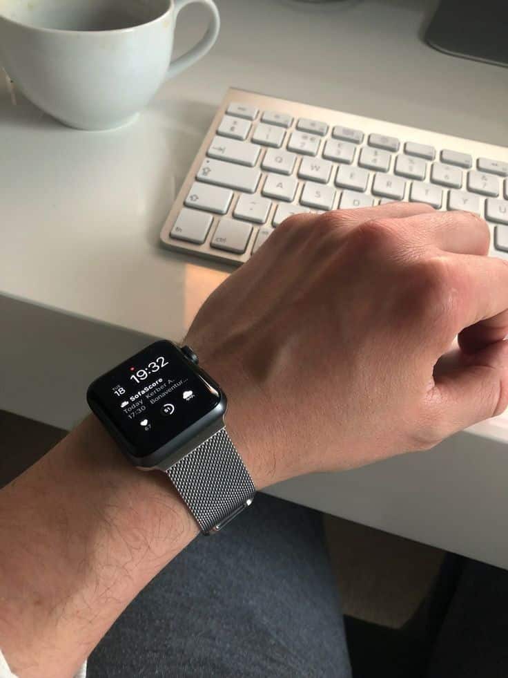 Person wearing a smartwatch with a mesh band, typing on a white keyboard, cup of coffee nearby on desk.