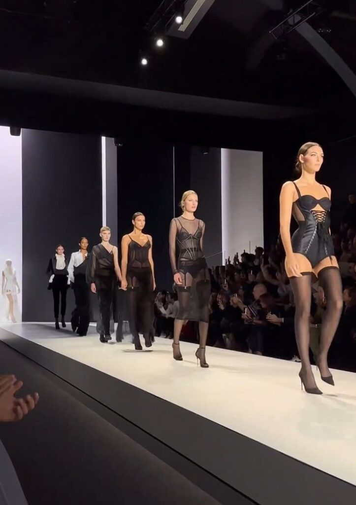 Models in stylish black outfits walk runway at fashion show, spotlighting modern design and elegance.