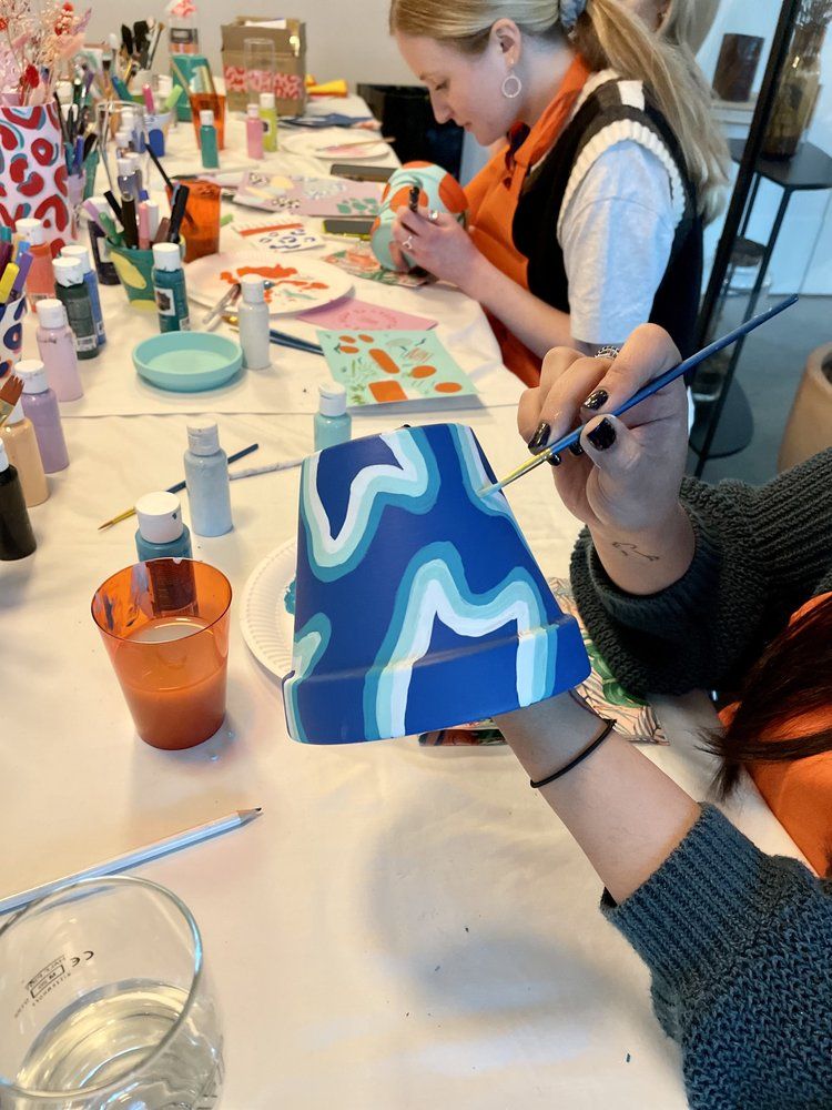 People painting colorful designs on pots at an art class, surrounded by brushes and paints.