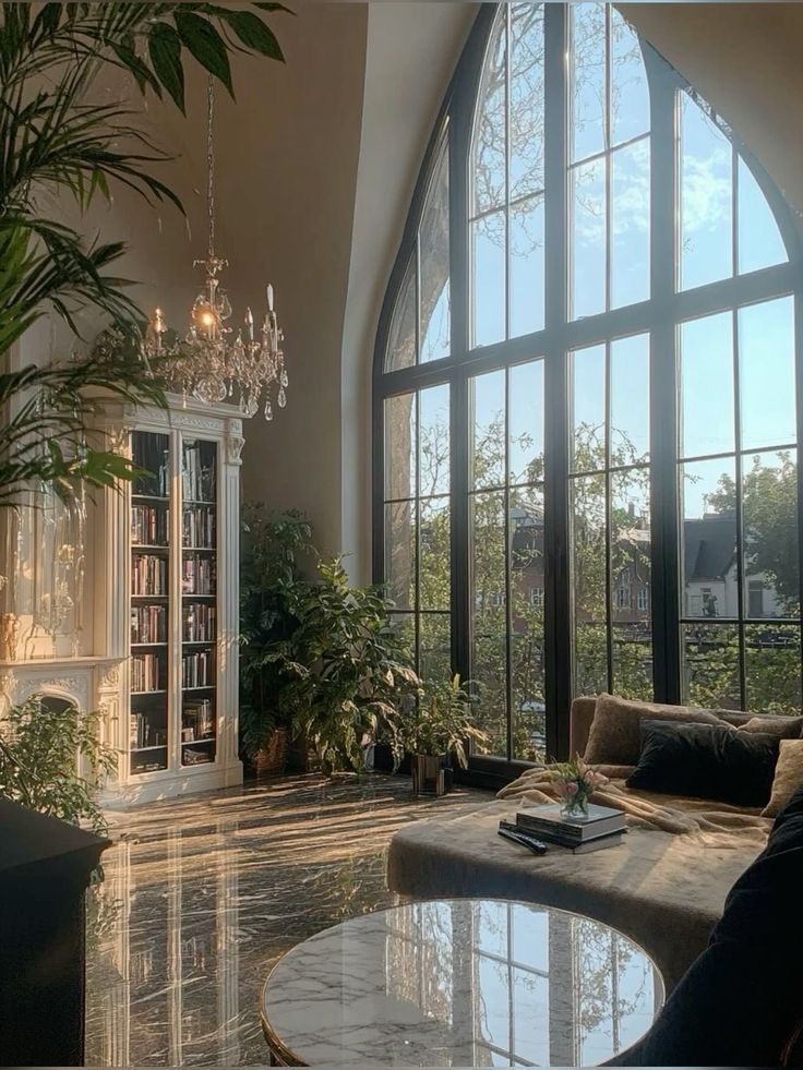 Luxurious living room with large arched windows, a chandelier, lush plants, cozy seating, and elegant decor.