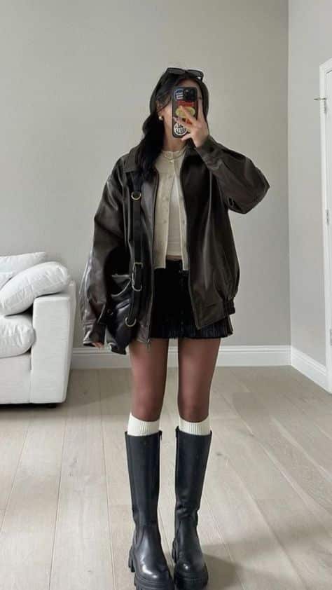 Person in stylish fall outfit with leather jacket, mini skirt, knee-high boots, and phone, standing in a modern living room.