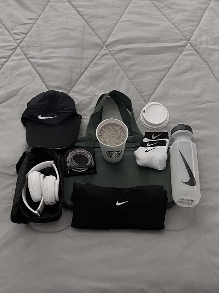 Black cap, Nike shirt, headphones, iced coffee, socks, and water bottle laid out for a workout on gray quilted background.