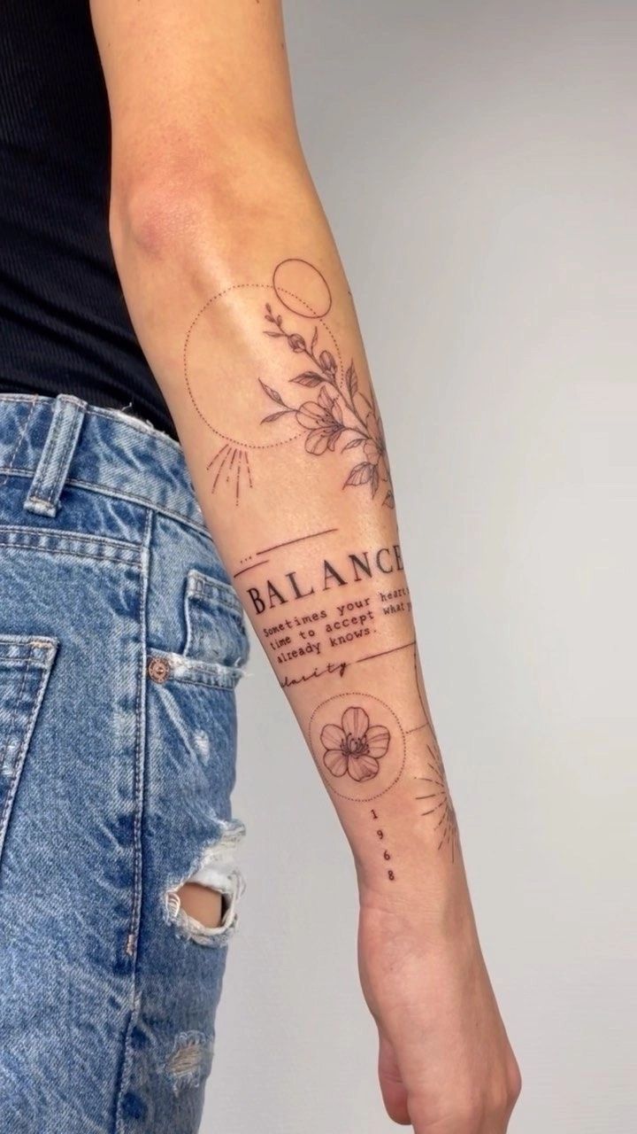 Forearm tattoo with flowers, geometric shapes, 'Balance' text, and meaningful words in black ink, worn with denim jeans.