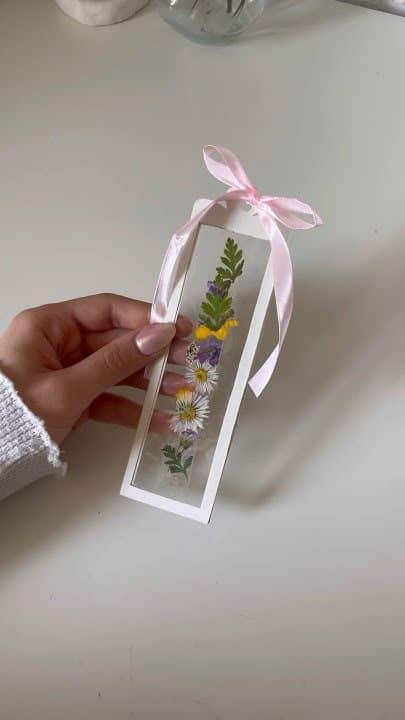 Hand holding a floral bookmark with pressed flowers and a pink ribbon on a white background.