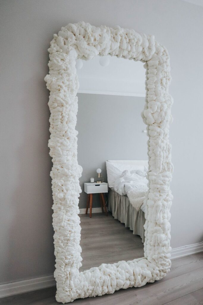Large rectangular mirror with textured white frame reflecting a cozy bedroom setting.