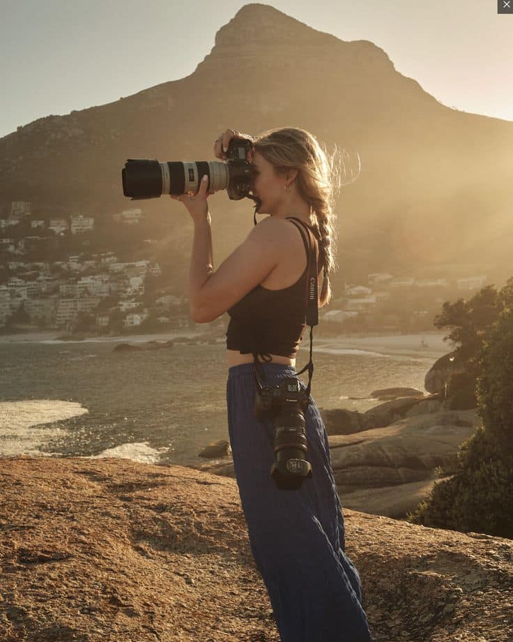 Photographer on a rocky cliff capturing a scenic coastal view at sunset with mountains in the background.