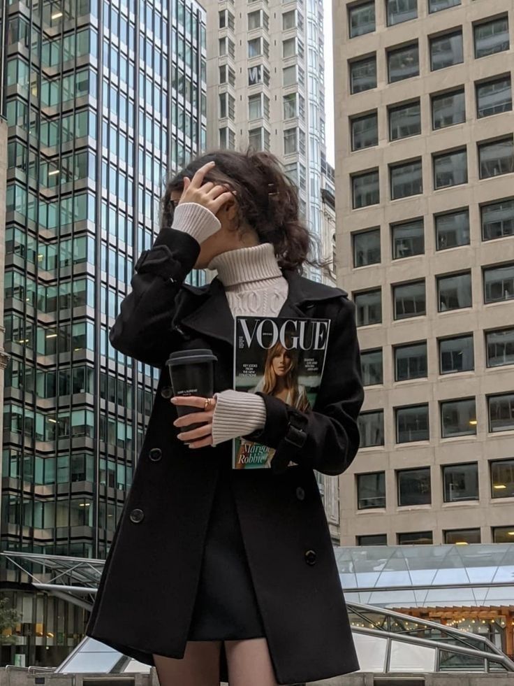 Woman in urban setting holding Vogue magazine and coffee cup, wearing stylish black coat and white turtleneck.