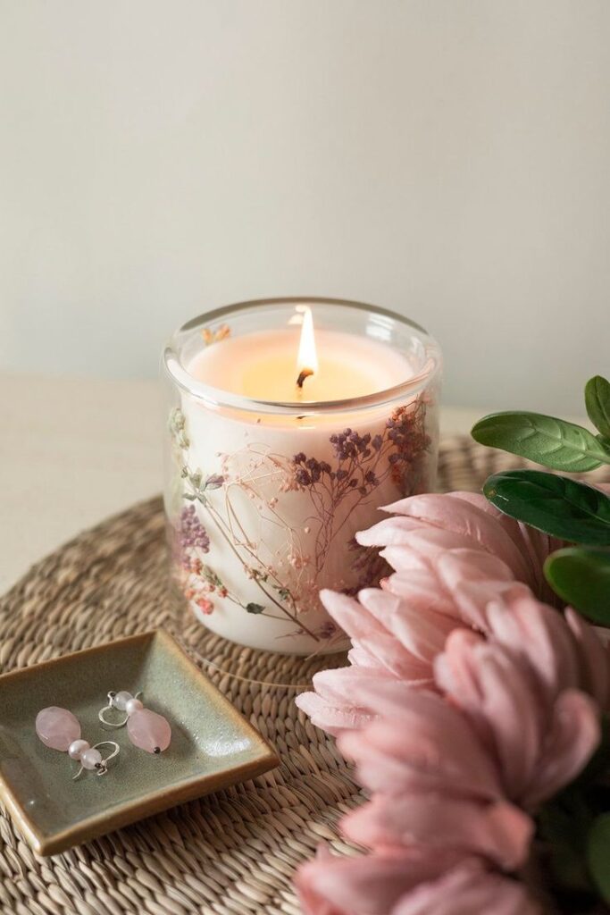 Lit floral candle beside pink flowers and earrings on a woven mat, creating a cozy, elegant ambiance.
