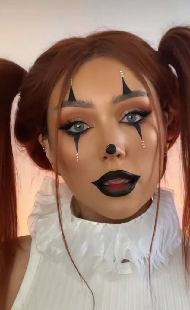 Person in clown makeup with dramatic black designs, white ruffled collar, and brown hair in pigtails.