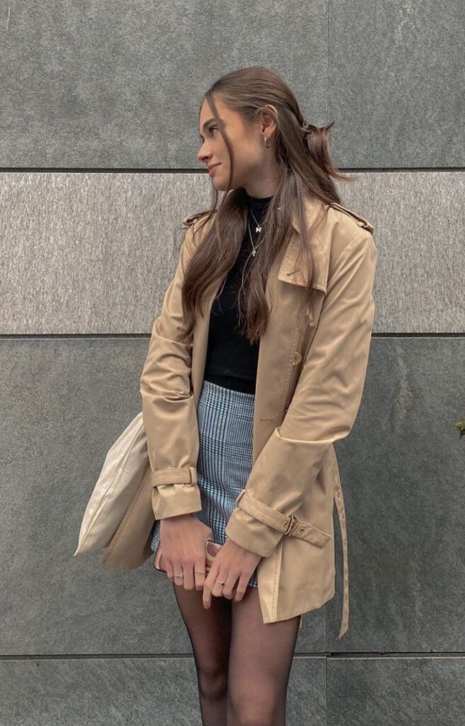 avery kylie grambs Woman in a beige trench coat posing against a gray wall, wearing a black top and plaid skirt, holding a bag. | Sky Rye Design Woman in a beige trench coat posing against a gray wall, wearing a black top and plaid skirt, holding a bag.