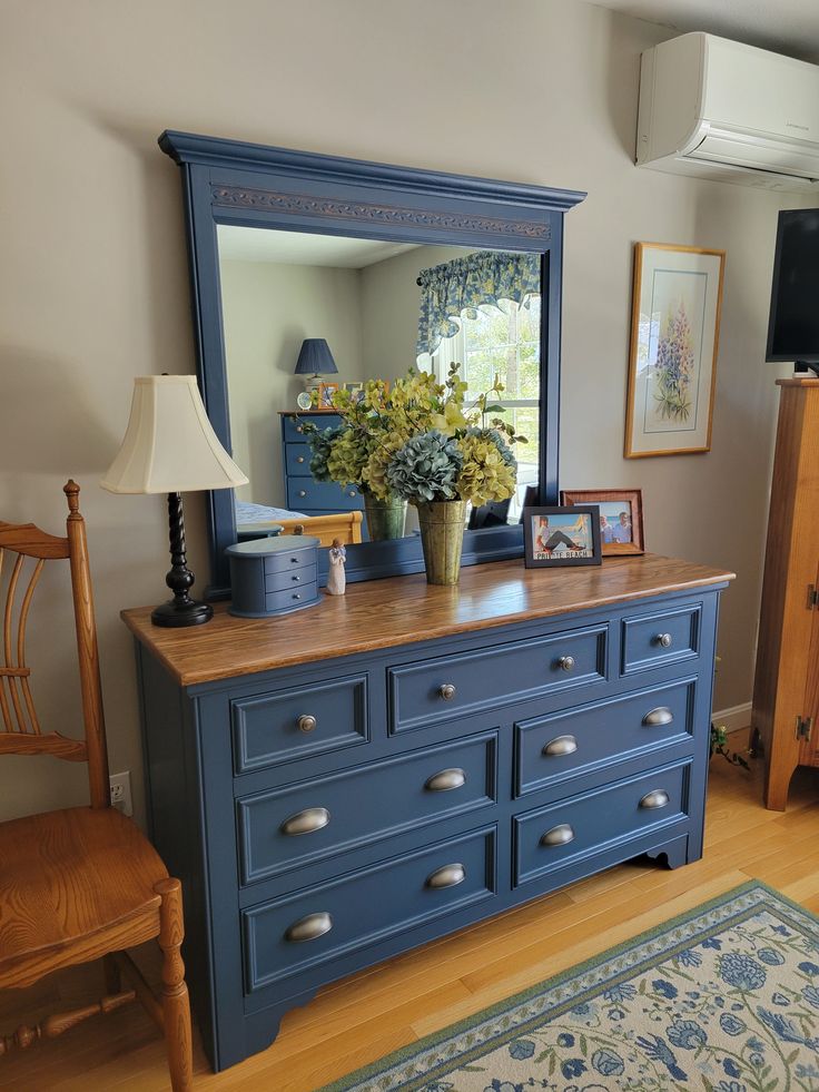 b0c6c64536983c23bdc745a18c6a691b Blue dresser with mirror, floral decor, and wooden top in cozy room with artwork and wooden chair nearby. | Sky Rye Design Blue dresser with mirror, floral decor, and wooden top in cozy room with artwork and wooden chair nearby.