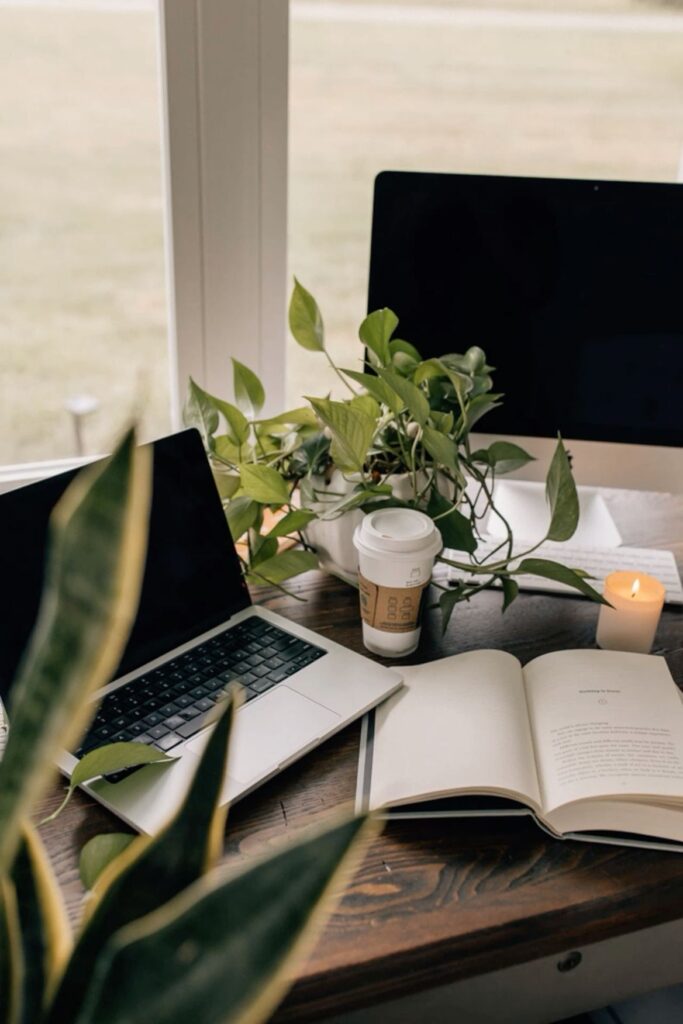 Cozy workspace with laptop, open book, coffee, candle, and houseplants on a wooden desk by a window.