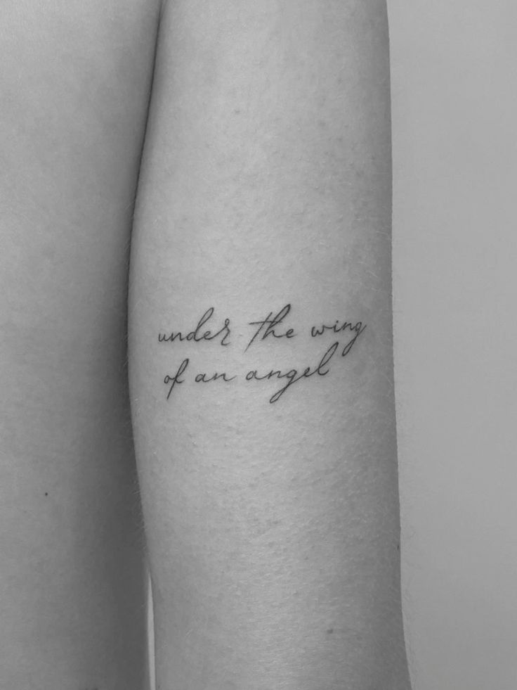 Minimalist script tattoo on arm reading under the wing of an angel in black ink, shown in close-up view.